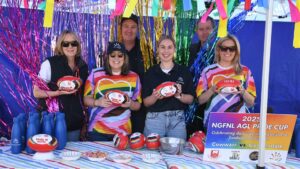 AGL Loy Yang team members attending the Pride Cup. Five people dressed in rainbow colours holding footballs standing at a stall surrounded by coloured streamers.