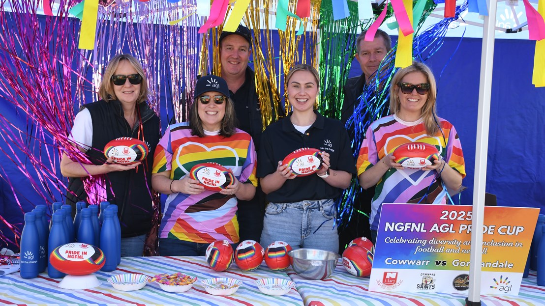 AGL Loy Yang team members attending the Pride Cup. Five people dressed in rainbow colours holding footballs standing at a stall surrounded by coloured streamers.