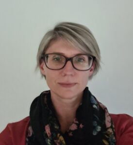 Image of Alexandra Lazarus-Priestley, has short blond hair and is wearing black rimmed glasses, a black scarf and a red top.