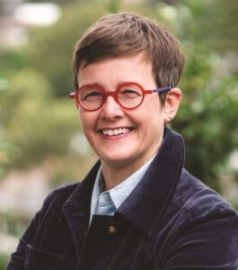 Dr Janin Bredehoeft, a person with short brown hair wearing glasses with red frames and a white shirt and blue jacket, smiling at the camera