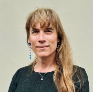 Image of Eloise Brook wearing a black top, and blue dangly earings. She has long blond hair and is smiling at the camera.
