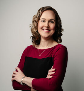Headshot of Marni Panas. A woman with short blond curly hair wearing a black and red top smiling at the camera.
