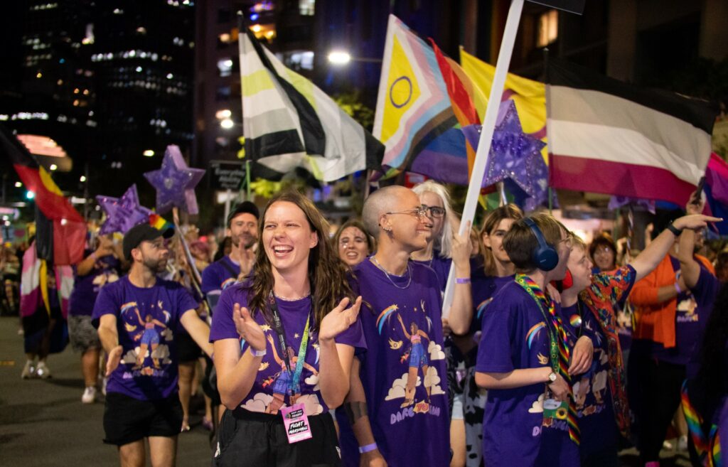 A group of people wearing purple shirts among a large crowd of people, many holding Pride flags.