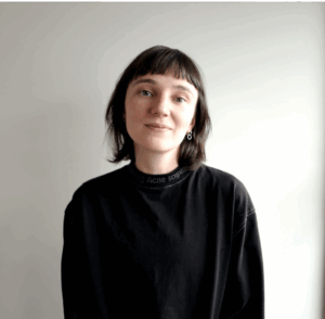 Charlie Farmer (she/they), LGBTQIA+ Education Presenter at Minus18. A person with shoulder length brown hair, wearing a black long sleeved top smiling