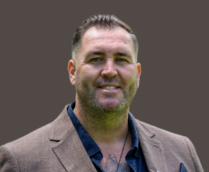 Image if Travis, wearing a brown blazer, nazy shirt and has short brown hair